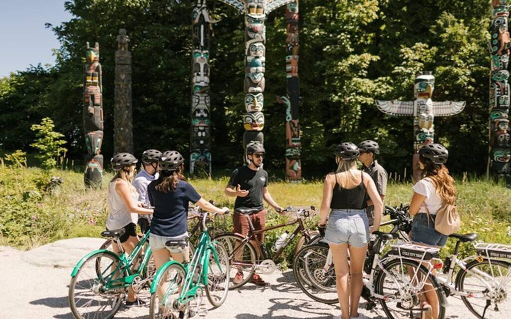a group stands in front of totem poles on the stanley park bike tour