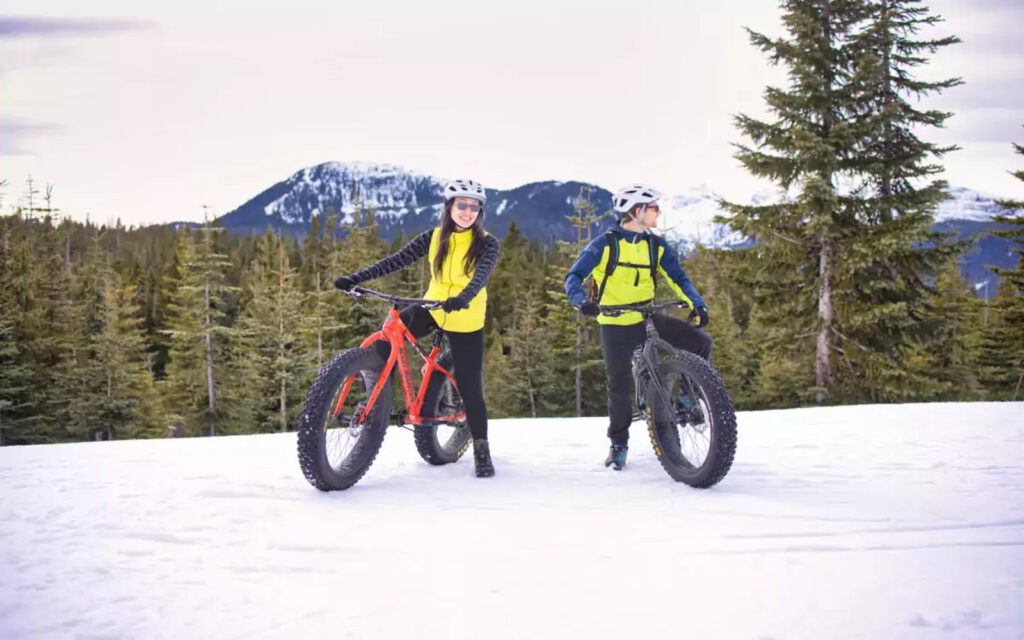 two people are fat biking in Vancouver during winter, wearing helmets, glasses, pants, and jackets