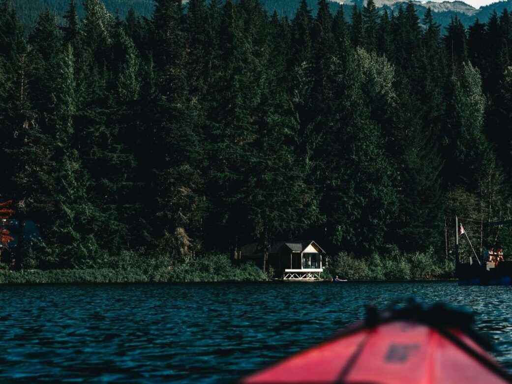 portion of a red paddleboard in the middle of the lake showing a lakehouse, green trees and dark blue water