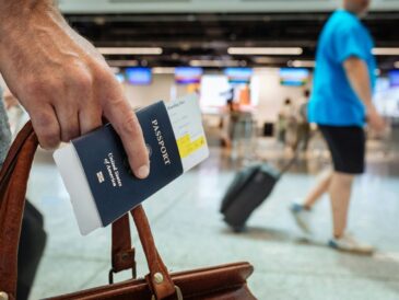 a man's hand holding a passport with the plane ticket inserted and a brown leather bag